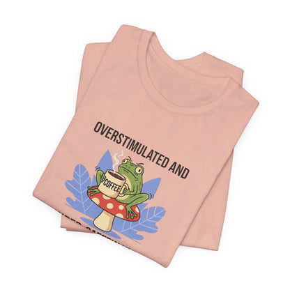 Peach t-shirt with a frog on a mushroom, reading "OVERSTIMULATED AND COFFEE".